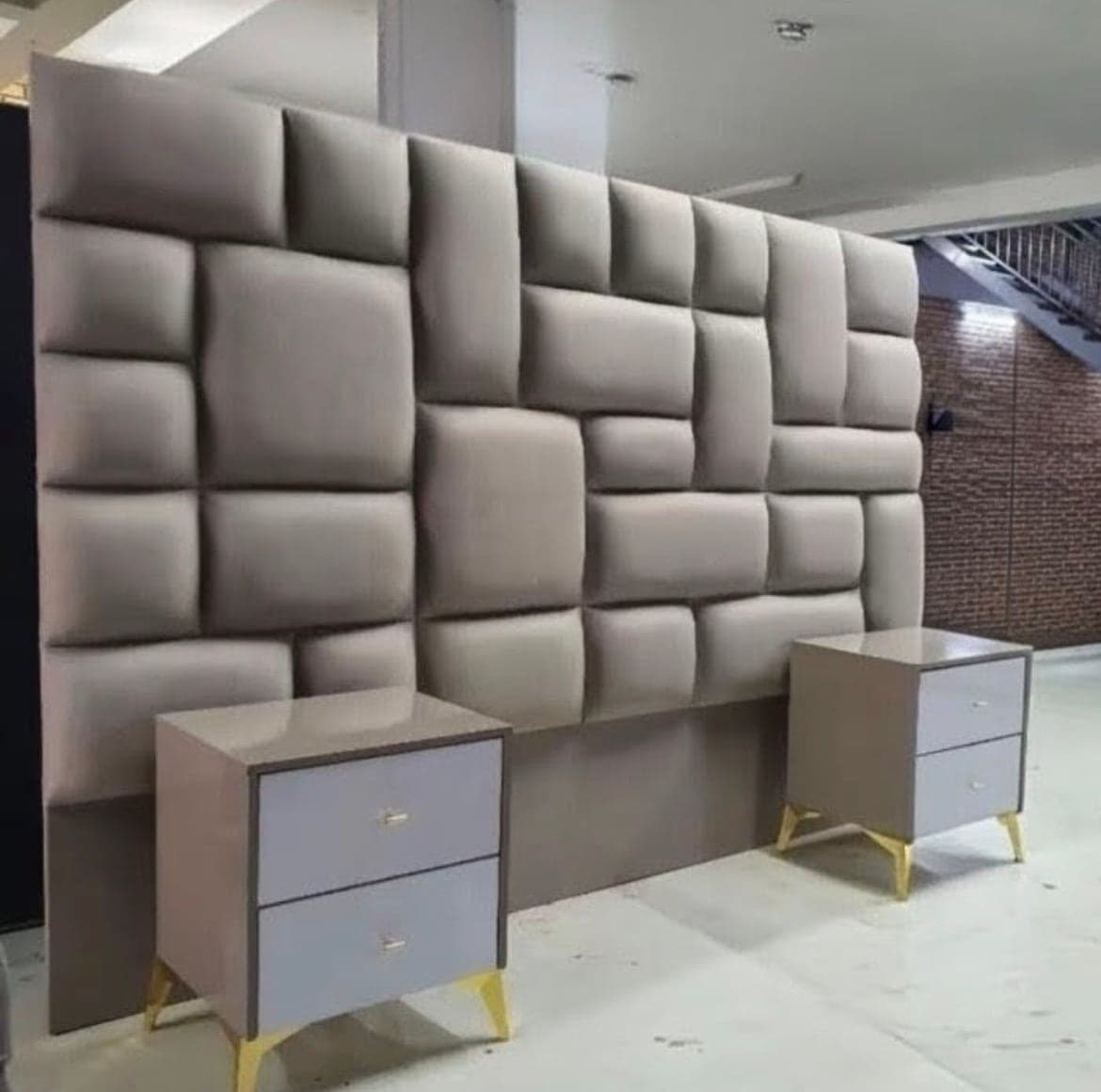 Taupe upholstered headboard of padded blocks in an offset grid, with a two-drawer pedestal on gold legs at each side.