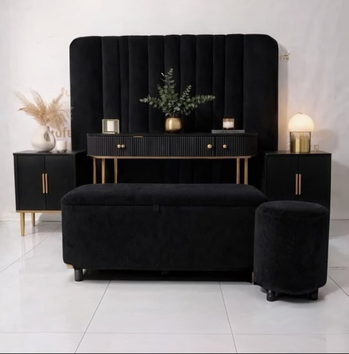 Black bedroom suite with a fluted velvet headboard, gold-trimmed console, two pedestals, a storage bench and a round ottoman.